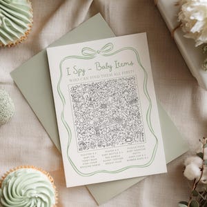 May include: A baby shower game card with the text "I Spy - Baby Items" and a detailed illustration of baby-related objects. The card is on a sage green envelope, with cupcakes and a wrapped gift in the background.