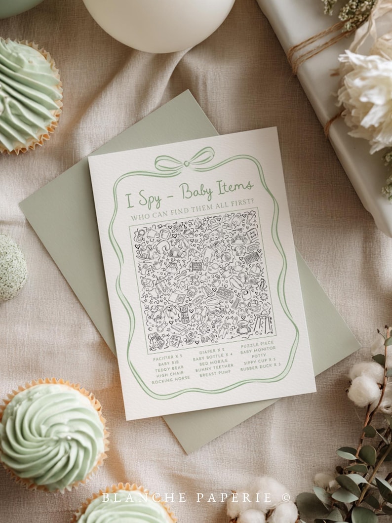 May include: A baby shower game card with the text "I Spy - Baby Items" and a detailed illustration of baby-related objects. The card is on a sage green envelope, with cupcakes and a wrapped gift in the background.