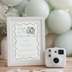 May include: A white framed sign with the text "Snap a Pic & Share the Love" and a white instant camera. The sign includes instructions for a baby shower activity. Soft green balloons and flowers are in the background.