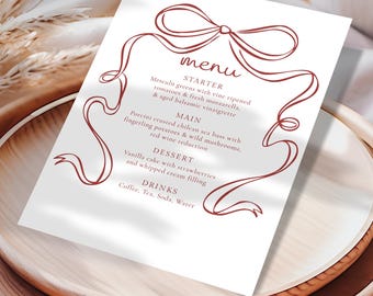 Op de afbeelding: Een witte menukaart met een bordeauxrood lintontwerp en tekst. Het menu bevat een voorgerecht, hoofdgerecht, dessert en drankjes. Het menu staat op een houten dienblad.