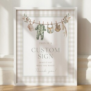May include: A framed sign with a watercolor design. A clothesline displays baby items: a cap, camouflage onesie, bib, boots, and antlers. The sign reads "THIS IS A CUSTOM SIGN" and "MAKE IT SAY WHAT YOU WANT!" with "Thank you!" below.