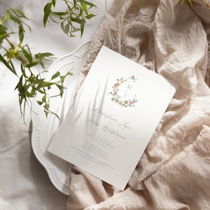May include: A wedding invitation with a floral wreath design and the names "Madeline Love" and "Allen Wilkinson" in calligraphy. The invitation details the date and time of the wedding, which is Saturday, July 1st, at 4:00 p.m. at the Padua Hills Theatre in Claremont, CA.