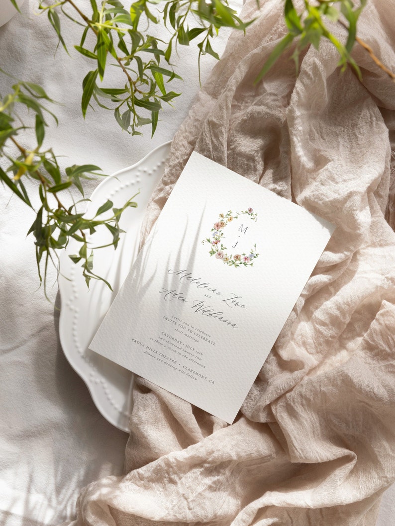 May include: A wedding invitation with a floral wreath design and the names "Madeline Love" and "Allen Wilkinson" in calligraphy. The invitation details the date and time of the wedding, which is Saturday, July 1st, at 4:00 p.m. at the Padua Hills Theatre in Claremont, CA.