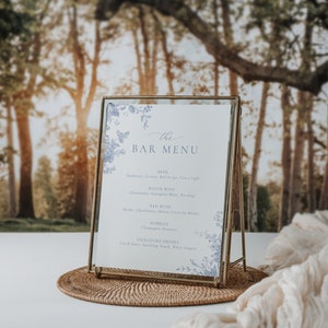 Pu&ograve; includere: Un cartello con cornice dorata, sfondo bianco e dettagli floreali blu. Il cartello recita "the BAR MENU" e elenca diverse opzioni di birra, vino e cocktail.