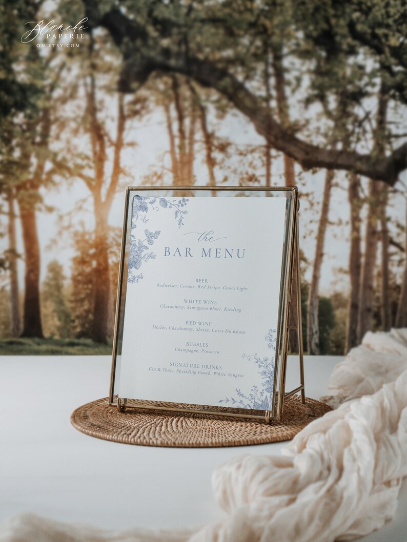 Pu&ograve; includere: Un cartello con cornice dorata, sfondo bianco e dettagli floreali blu. Il cartello recita "the BAR MENU" e elenca diverse opzioni di birra, vino e cocktail.