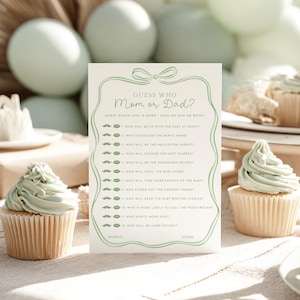 May include: A baby shower game card with the text "Guess Who Mom or Dad?" in a green frame, with questions about the parents. Two cupcakes with green frosting are in the foreground, with balloons in the background.