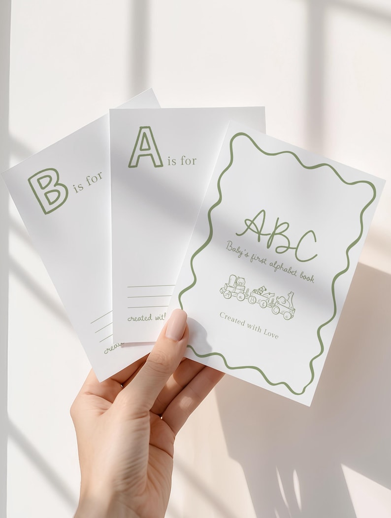May include: A set of white ABC flashcards with green lettering and illustrations. The cards are held in a hand and feature the letters A and B, along with the text "Baby's first alphabet book" and "Created with Love."