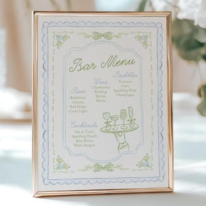 May include: A framed bar menu with a gold-colored frame. The menu features a light blue and white design with green text. The menu lists beer, wine, and cocktail options, including Gin & Tonic and Sparkling Punch.