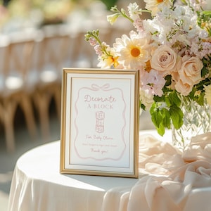 Pu&ograve; includere: Un cartello incorniciato con bordo dorato e sfondo bianco. Il cartello ha un testo rosa che recita "Decorate a Block" e "For Baby Olivia". Un bouquet di fiori &egrave; posto dietro il cartello. Il tavolo &egrave; coperto da una tovaglia color crema.