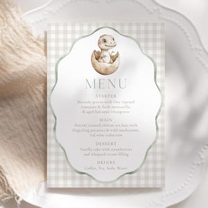 Può includere: Un biglietto del menu con uno sfondo a quadretti vichy verde chiaro e bianco. Il menu presenta un'illustrazione ad acquerello di un cucciolo di dinosauro che esce da un uovo. La parola "MENU" è in alto, con le voci del menu elencate di seguito.