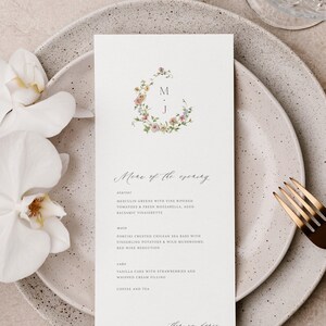 May include: A wedding menu card with a floral wreath design and the initials "M.J." in the center. The menu includes a starter, main course, cake, and coffee and tea. The text "Menu of the evening" is written in cursive at the top of the card.