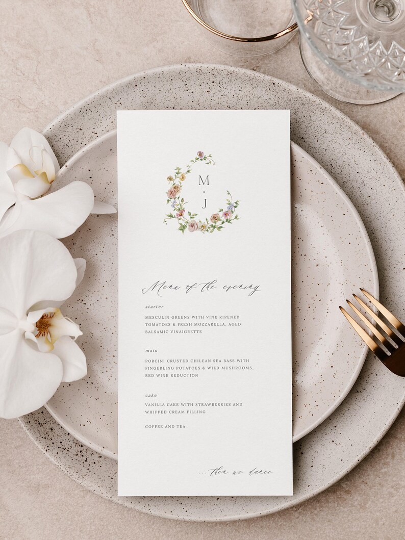 May include: A wedding menu card with a floral wreath design and the initials "M.J." in the center. The menu includes a starter, main course, cake, and coffee and tea. The text "Menu of the evening" is written in cursive at the top of the card.