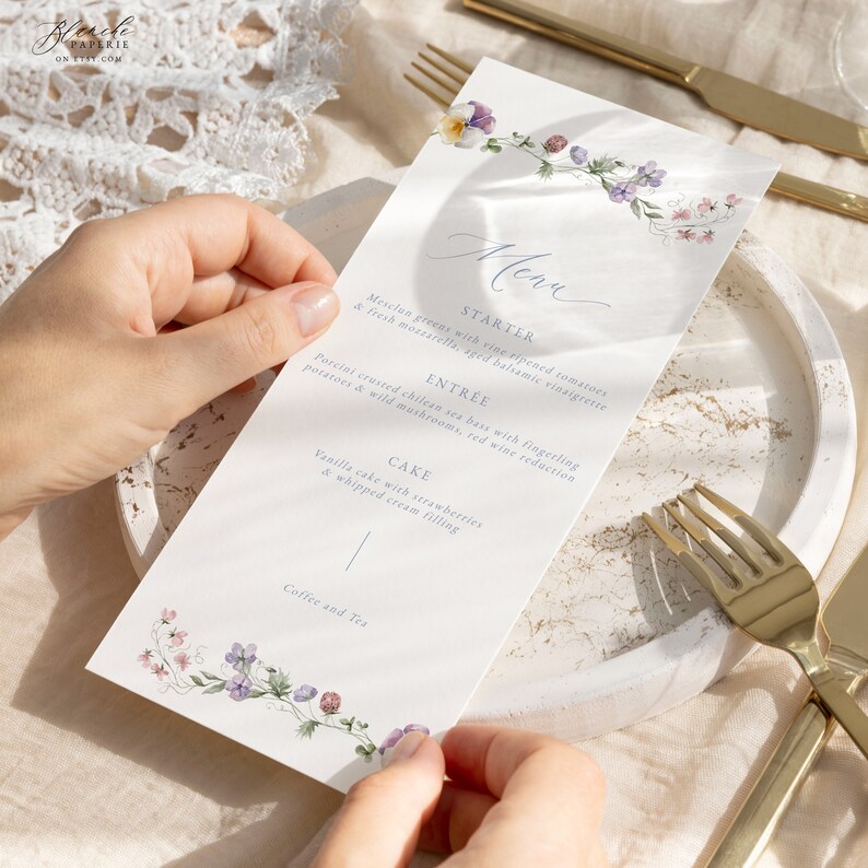 May include: A white menu card with a floral border and handwritten text. The menu includes a starter, entree, cake, and coffee and tea. The text reads: "Menu, Starter, Mesclun greens with vine ripened tomatoes & fresh mozzarella, aged balsamic vinaigrette, Entree, Porcini crusted chilean sea bass with fingerling potatoes & wild mushrooms, red wine reduction, Cake, Vanilla cake with strawberries & whipped cream filling, Coffee and Tea."