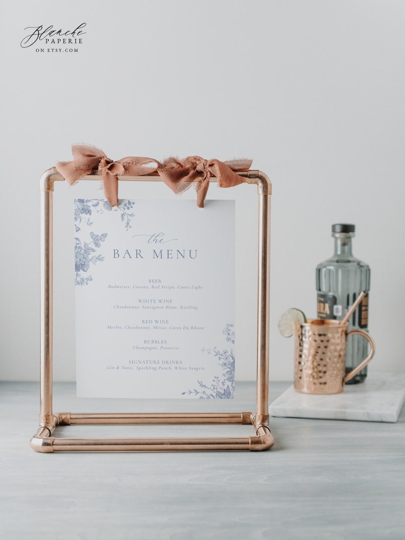 Pu&ograve; includere: Un supporto in tubo di rame con un cartoncino bianco con un motivo floreale e il testo "the BAR MENU". Il cartoncino elenca diversi tipi di birra, vino bianco, vino rosso, bollicine e bevande firmate.