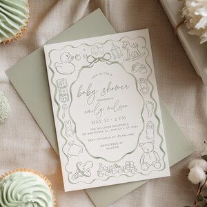 May include: Baby shower invitation with a sage green envelope and a matching invitation card. The card features a hand-drawn border of baby-themed illustrations and the text "Join us for a baby shower honoring Emily Wilson". Two cupcakes are visible.