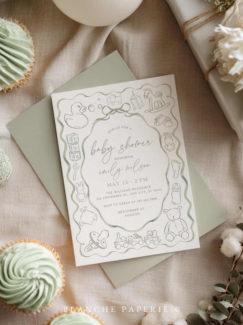 May include: Baby shower invitation with a sage green envelope and a matching invitation card. The card features a hand-drawn border of baby-themed illustrations and the text "Join us for a baby shower honoring Emily Wilson". Two cupcakes are visible.
