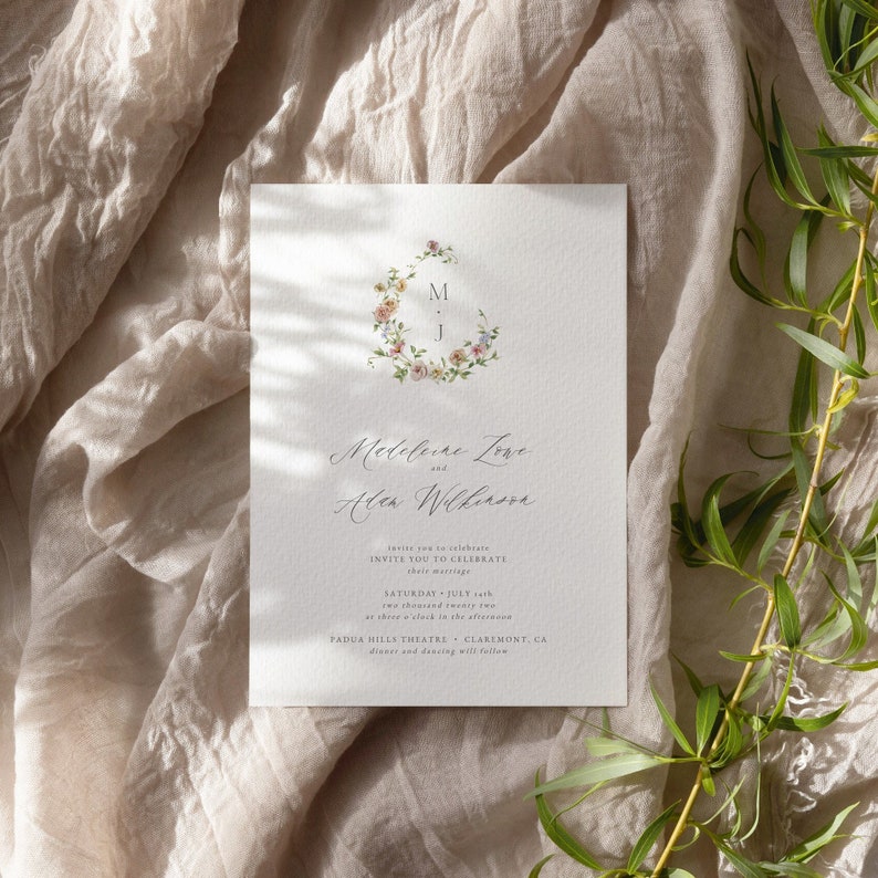 May include: Wedding invitation with a floral wreath design featuring the initials M and J. The invitation is for Medeleine Love and Alen Wilkinson, and it states the wedding will take place on Saturday, July 22nd at three o'clock in the afternoon at Padua Hills Theatre in Claremont, CA. Dinner and dancing will follow.