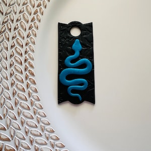 May include: A black and blue snake charm with a textured background. The charm is shaped like a rectangle with rounded corners and has a hole at the top for hanging.