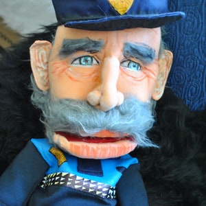 Op de afbeelding: Een blauwe en grijze pop met een blauwe politiehoed en een grijze snor. De pop draagt een blauw shirt met een zwart-witte gepatenteerde riem.
