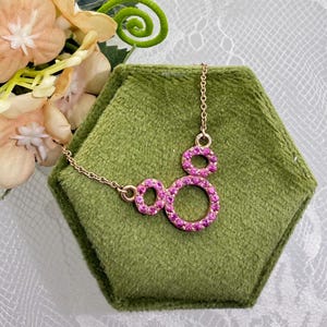 May include: A gold-toned necklace with a pendant featuring three connected circles, each encrusted with small, vibrant pink gemstones. The necklace rests on a green velvet hexagonal display, with floral accents in the background.