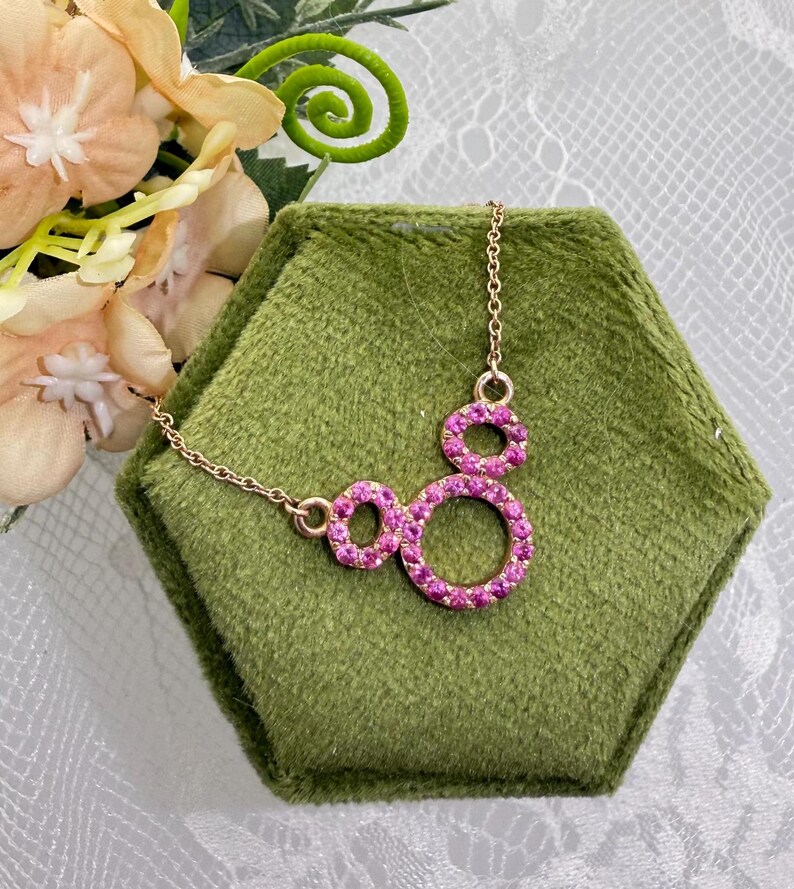 May include: A gold-toned necklace with a pendant featuring three connected circles, each encrusted with small, vibrant pink gemstones. The necklace rests on a green velvet hexagonal display, with floral accents in the background.