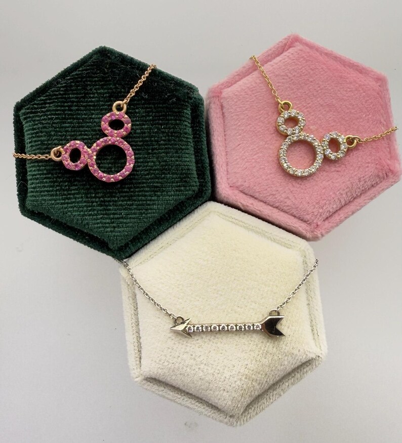 May include: Three necklaces displayed on velvet boxes. One gold necklace features a Mickey Mouse-shaped pendant with pink gemstones. Another gold necklace has a similar pendant with clear gemstones. The third necklace is silver with an arrow pendant.