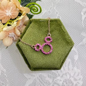 May include: A gold-toned necklace with a pendant featuring three connected circles, each encrusted with pink gemstones. The necklace is displayed on a green hexagonal surface, with flowers in the background.