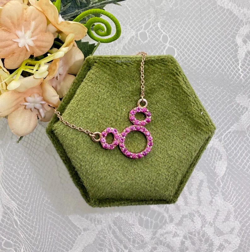 May include: A gold-toned necklace with a pendant featuring three connected circles, each encrusted with pink gemstones. The necklace is displayed on a green hexagonal surface, with flowers in the background.