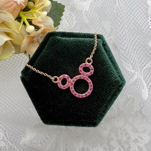 May include: A gold-colored necklace with a pendant featuring three connected circles, each outlined with pink gemstones. The necklace is displayed on a dark green velvet surface, with a floral arrangement in the background.