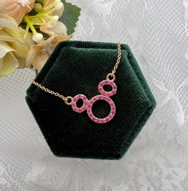 May include: A gold-colored necklace with a pendant featuring three connected circles, each outlined with pink gemstones. The necklace is displayed on a dark green velvet surface, with a floral arrangement in the background.