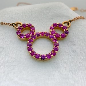 May include: A gold-toned necklace featuring a pendant in the shape of Mickey Mouse's head. The pendant and chain are gold. The Mickey Mouse head is outlined with a ring of small, bright pink gemstones.