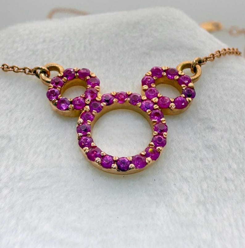 May include: A gold-toned necklace featuring a pendant in the shape of Mickey Mouse's head. The pendant and chain are gold. The Mickey Mouse head is outlined with a ring of small, bright pink gemstones.