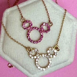 May include: Two gold-toned necklaces with Mickey Mouse pendant designs. One pendant is encrusted with pink gemstones, the other with clear gemstones. The necklaces rest on a white surface against a pink background.