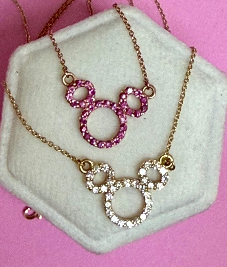 May include: Two gold-toned necklaces with Mickey Mouse pendant designs. One pendant is encrusted with pink gemstones, the other with clear gemstones. The necklaces rest on a white surface against a pink background.