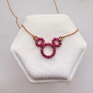 May include: A rose gold necklace featuring a Mickey Mouse-shaped pendant. The pendant is encrusted with small, vibrant pink gemstones. The necklace is displayed on a white, hexagonal surface.