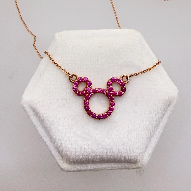 May include: A rose gold necklace featuring a Mickey Mouse-shaped pendant. The pendant is encrusted with small, vibrant pink gemstones. The necklace is displayed on a white, hexagonal surface.