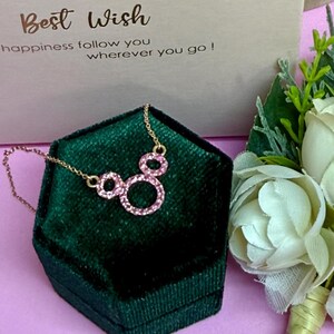 May include: A gold-colored necklace with a pendant of three pink circles, resting on a dark green velvet jewelry box. The box is set against a light pink surface, with a card that reads "Best Wish" and a bouquet of white flowers.