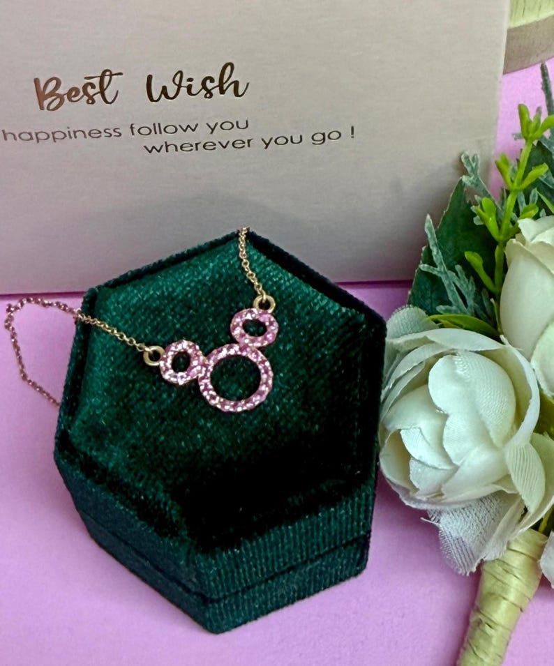 May include: A gold-colored necklace with a pendant of three pink circles, resting on a dark green velvet jewelry box. The box is set against a light pink surface, with a card that reads "Best Wish" and a bouquet of white flowers.