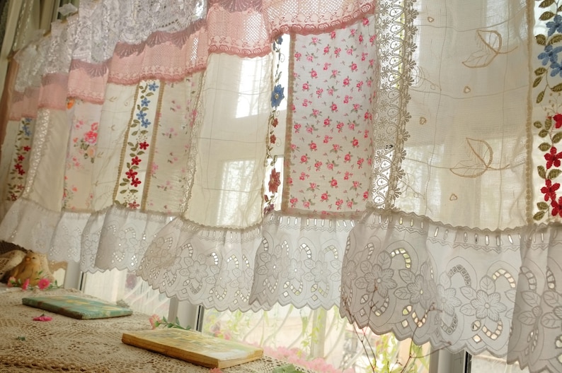 May include: A white sheer curtain with a lace trim and patchwork design. The curtain is made of different fabrics with floral patterns and embroidery. The curtain is hung on a window with sunlight shining through.