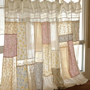 May include: A patchwork curtain made from various fabrics, including floral patterns, lace, and plain materials. The curtain features a mix of pink, yellow, and white, with a ruffled hem and lace detailing. It hangs from a wooden pole.