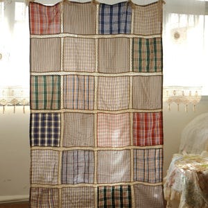 May include: A patchwork curtain with a variety of plaid patterns in red, blue, green, and brown. The curtain is made of squares sewn together with a decorative trim. The curtain hangs from a rod with knotted ties.