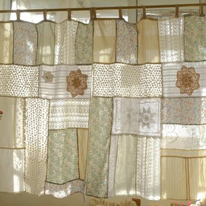 Puede incluir: Una cortina de patchwork con una variedad de telas, incluyendo estampados florales, a rayas y de encaje. La cortina presenta una mezcla de colores crema, beige y verde claro, con elementos decorativos de ganchillo. La cortina cuelga de una barra de madera.