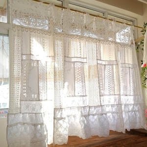 Puede incluir: Cortinas de encaje blanco colgadas frente a una ventana. Las cortinas están hechas de varios patrones de encaje, creando un efecto en capas. La luz del sol se filtra a través de la delicada tela, iluminando la habitación. Tratamiento de ventana decorativo.