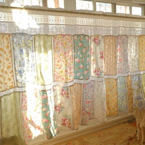 Könnte beinhalten: Ein Patchwork-Vorhang mit Blumenmustern in verschiedenen Farben, darunter Gelb, Blau und Rosa. Der Vorhang besteht aus mehreren Paneelen mit Spitzensaum und hängt vor einem Fenster, wodurch eine weiche, romantische Ästhetik entsteht.