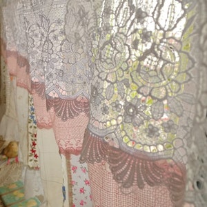 May include: White and pink lace curtains with a floral pattern. The curtains are hung on a window and are letting in light.