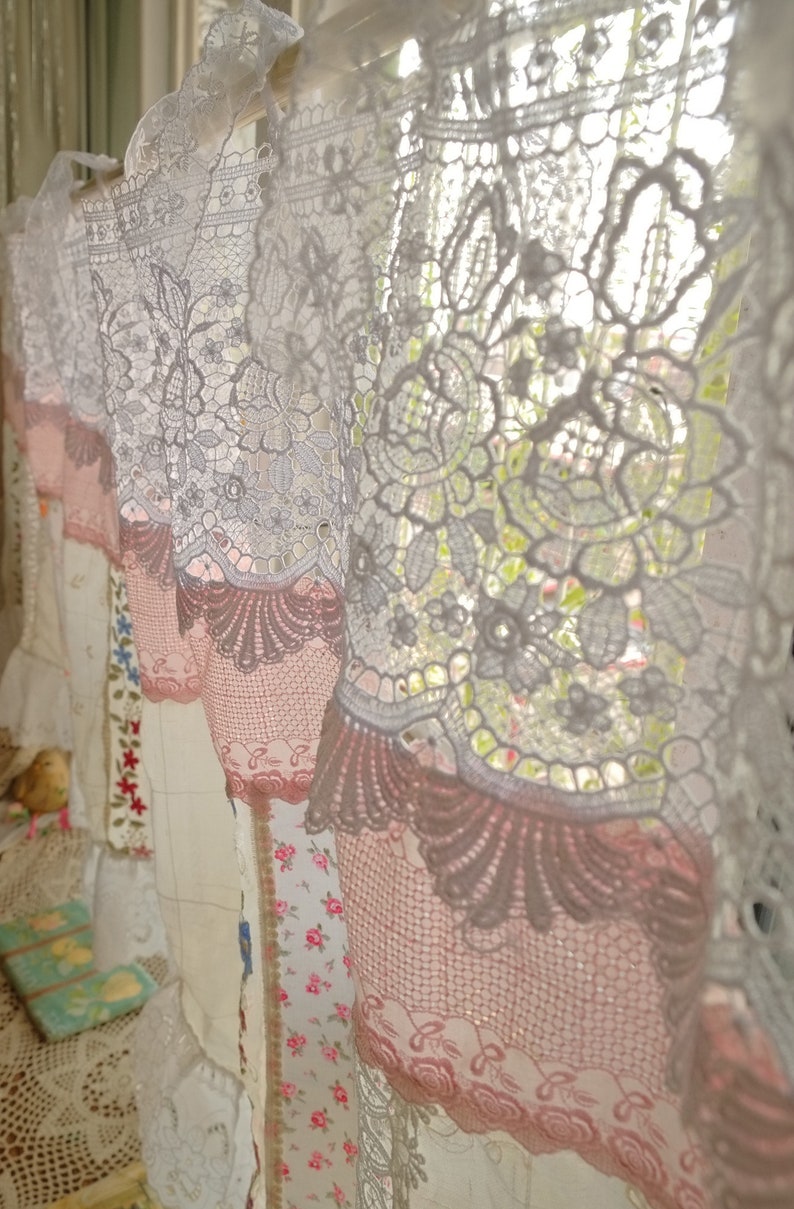 May include: White and pink lace curtains with a floral pattern. The curtains are hung on a window and are letting in light.