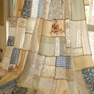 May include: A patchwork curtain with a variety of fabrics, including floral, gingham, and lace patterns. The curtain is a mix of beige, blue, and white, creating a vintage, handcrafted aesthetic. The curtain hangs from a wooden rod.