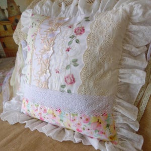 Könnte beinhalten: Ein quadratisches Zierkissen mit einem Patchwork-Design aus weißen, cremefarbenen und floralen Stoffen. Die Vorderseite zeigt vertikale Bahnen mit Rosenstickerei, Spitze und Lochstickerei, abgerundet durch einen breiten weißen Rüschenrand.