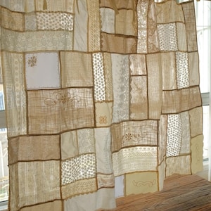 Pode incluir: Uma cortina de patchwork feita de vários painéis de tecido retangulares em tons de creme, bege e branco sujo. A cortina apresenta uma mistura de texturas, incluindo renda, linho bordado e algodão tecido, suspensa por uma vara de madeira com laços de tecido.