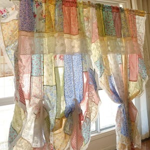 Puede incluir: Una cortina de patchwork con una variedad de telas en colores pastel, incluyendo estampados florales, cuadros vichy y lisos. La cortina está adornada con encaje y atada con lazos de tela, creando un aspecto en capas y texturizado. La cortina cuelga frente a una ventana.